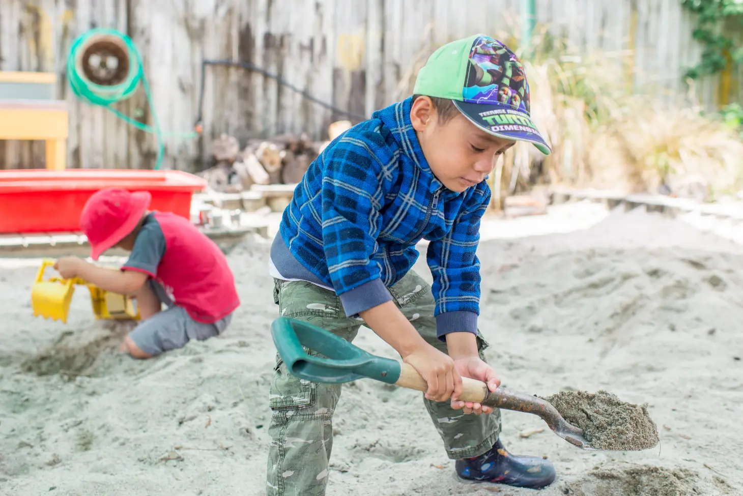 Kindergarten boy in green cap playing with a spade in a sandpit